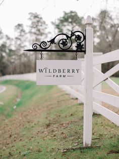 a sign that is hanging from the side of a white fence in front of a field