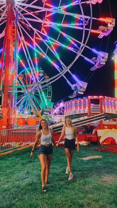 two women walking in front of a ferris wheel at night with colorful lights on it