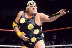 a man in a wrestling costume standing next to a ring holding a stick and ball