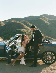 a man and woman sitting on the back of a car next to each other in front of a mountain