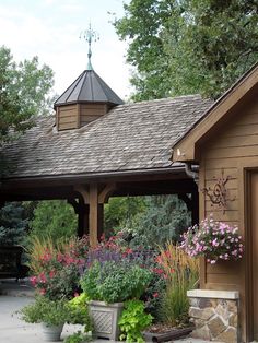 a gazebo surrounded by flowers and plants in front of a wooden building with a cupola on the roof