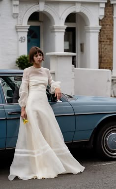 a woman in a white dress standing next to a blue car with her hand on the door handle