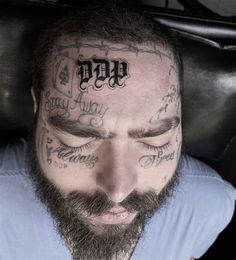 a bearded man with tattoos on his forehead and head looking down at the camera while wearing a blue shirt