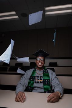 a man sitting at a desk with papers flying in the air behind him and wearing a graduation cap