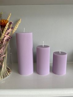 three purple candles sitting on top of a white shelf next to flowers and a vase