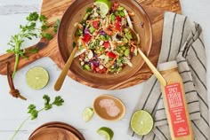 a wooden bowl filled with salad next to limes and seasoning on a cutting board