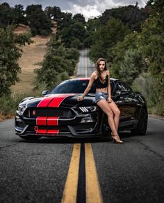 a woman sitting on the hood of a sports car in front of a road with trees