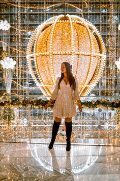 a woman standing in front of a giant christmas ball with lights all around her and wearing black boots