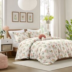 a white bed with pink flowers on it in a room next to a window and potted plant