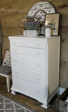 a white dresser sitting next to a wooden wall with a clock on it's face