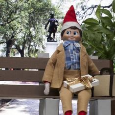 an elf doll sitting on a bench with a gift box in his hand and a statue behind it