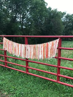 an orange and white striped cloth hanging from a red fence in a grassy field next to trees