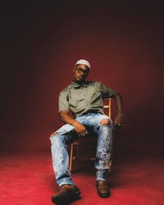 a man sitting on top of a wooden chair in front of a red wall with his legs crossed