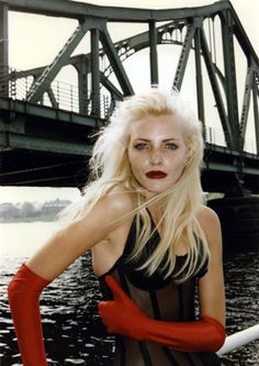 a woman with white hair and red gloves posing in front of a bridge over water