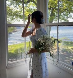 a woman standing in front of a window holding a bouquet of flowers and looking out the window