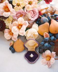 an assortment of fruit and flowers are displayed on a table with a ring box in the middle