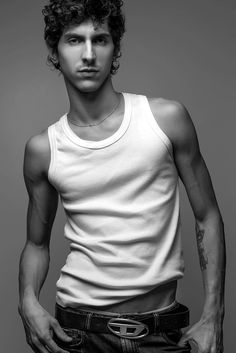 a man with curly hair wearing a white tank top and black jeans is posing for the camera