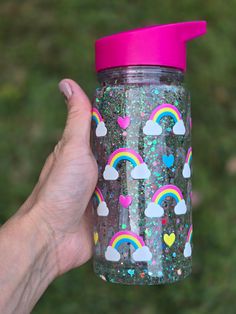 a hand holding a pink and purple glittered cup with rainbows, hearts, and clouds on it