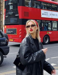 a woman in black jacket and sunglasses standing next to a red double decker bus