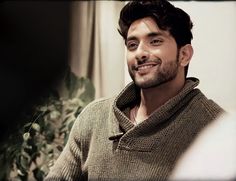 a man smiling and wearing a sweater in front of a potted plant with green leaves