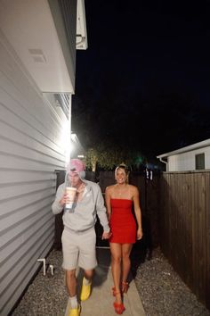 a man and woman walking down a sidewalk at night with one holding a drink in her hand