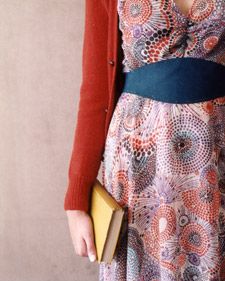 a woman in a dress and cardigan holding a yellow purse standing next to a wall