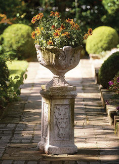 a stone urn with flowers in it sitting on a brick path surrounded by bushes