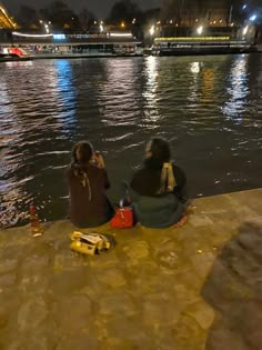 two people sitting on the edge of a body of water at night with boats in the background