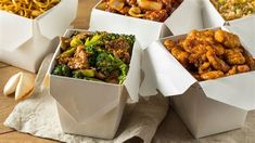 four white boxes filled with different types of food on top of a wooden table next to onion rings