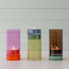 three different colored candles sitting next to each other on a white counter top in front of a wall