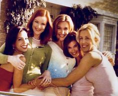 four women are posing together in front of a house and one woman is holding her arm around the other