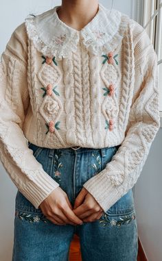 a woman wearing jeans and a sweater with flowers on the shoulders is standing in front of a window