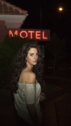 a woman with long hair standing in front of a motel sign at night, looking off to the side