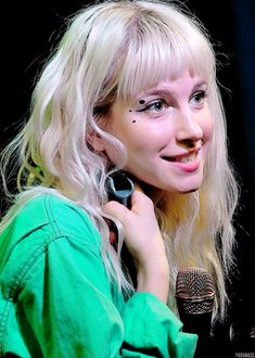 a woman with long blonde hair holding a microphone and talking on the phone while standing in front of a black background