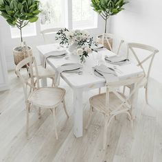 a dining room table set with white chairs and flowers in vases on the windowsill