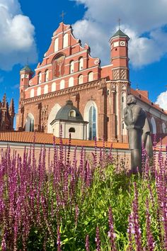 an old building with purple flowers in the foreground and a statue of a horse