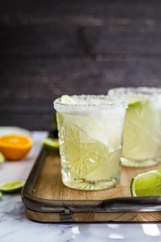 two margaritas on a cutting board with lime and an orange