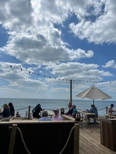 people are sitting at tables on the deck near the ocean and an umbrella is hanging over them