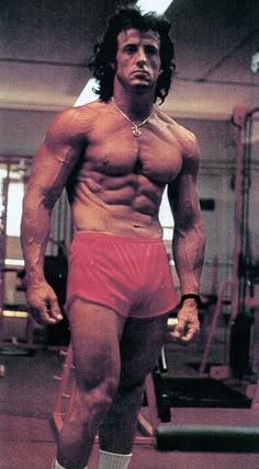 a shirtless man in red trunks and socks standing next to a gym equipment rack