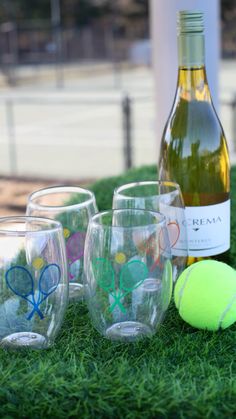 four wine glasses and a tennis ball on the grass next to a bottle of wine