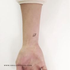 a small saturn tattoo on the left wrist is shown in black ink, and it has a tiny space ship at the center