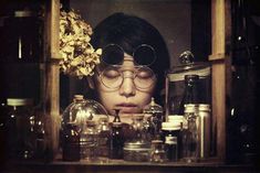 a woman with glasses and flowers in her hair behind a shelf full of glass jars