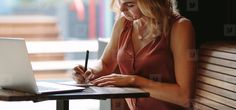 a woman sitting at a table with a laptop and pen in her hand, writing on the paper