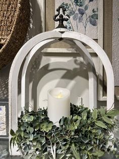 a candle is lit in front of a white arch with greenery around it on a table