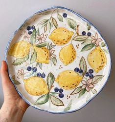 a hand holding a plate with lemons and blueberries painted on the side, in front of a gray wall