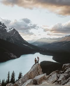 two people sitting on top of a large rock next to a body of water with mountains in the background