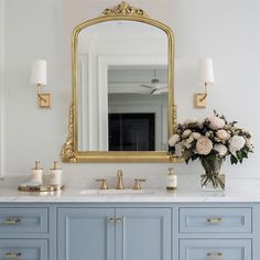 a bathroom vanity with blue cabinets and gold mirror above it, flowers in vase on the counter