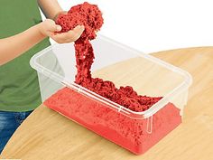 a child is playing with red sand in a plastic container on a wooden table,