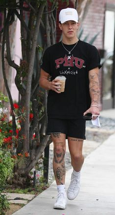 a man with tattoos walking down the street holding a drink and wearing a white hat