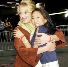 two women hugging each other in front of a fence and building at night with people behind them